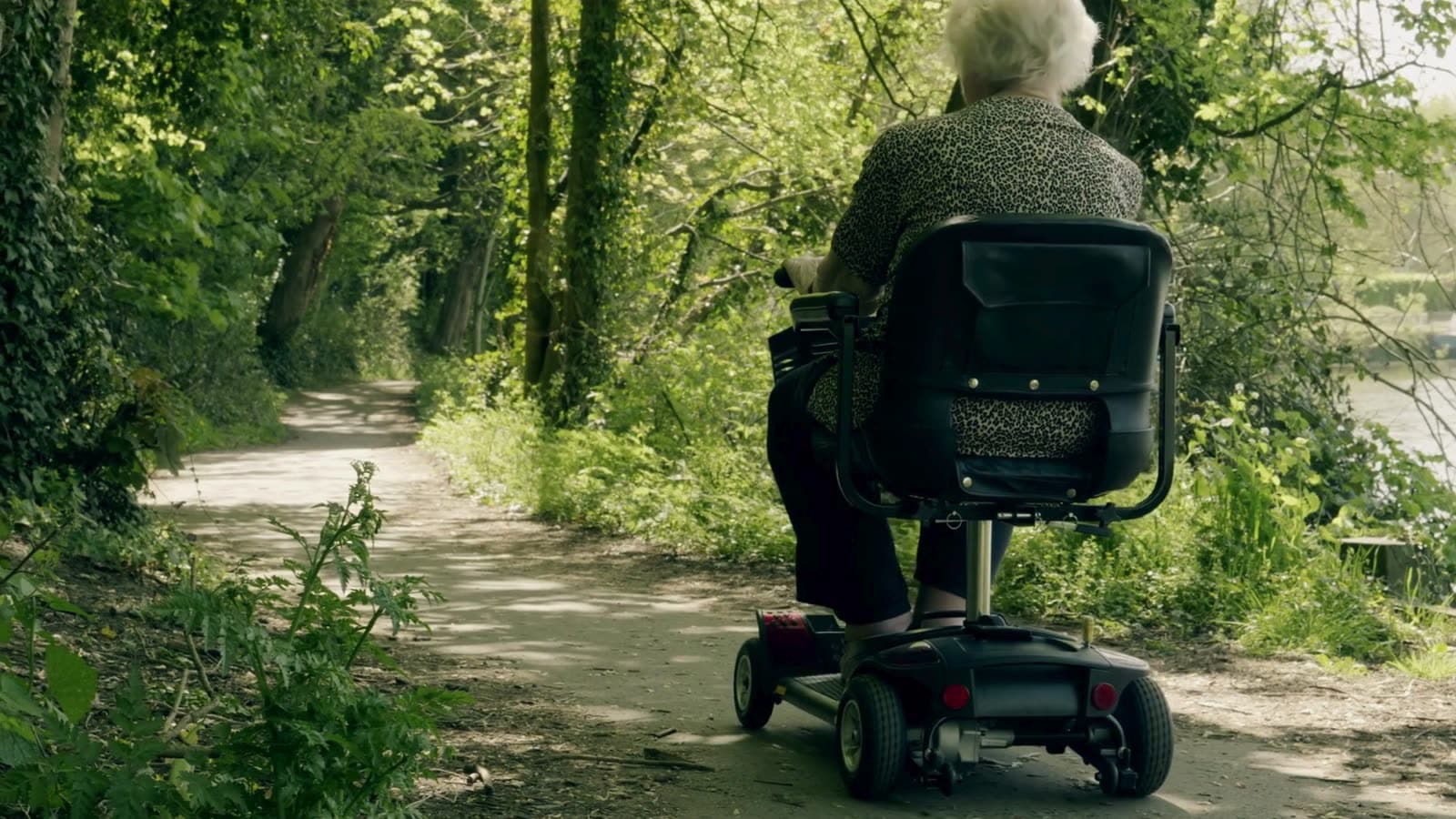A woman riding a mobility scooter along a tree-lined path on a bright summer day